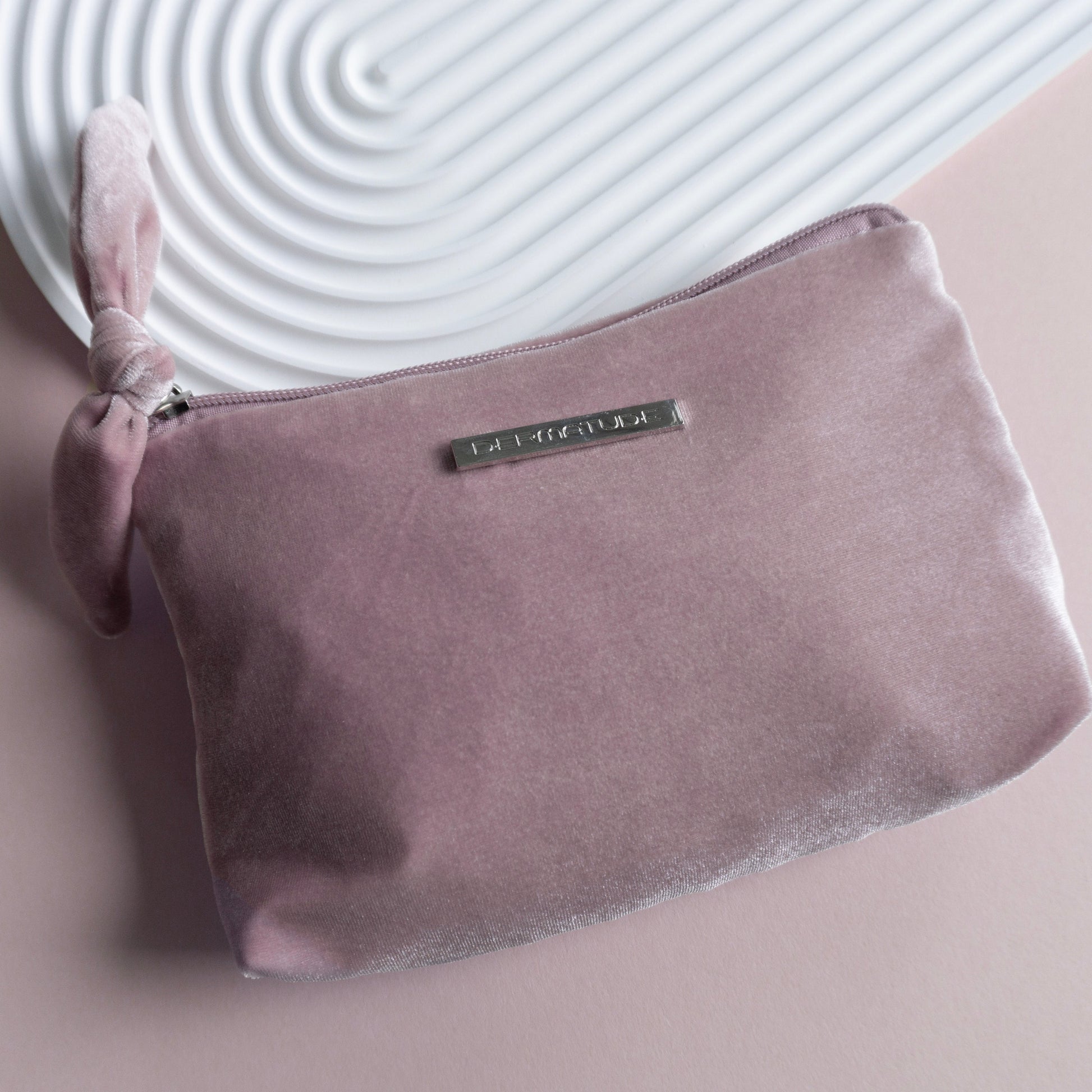 A pink velvet clutch with a silver pin that says DERMATUDE.