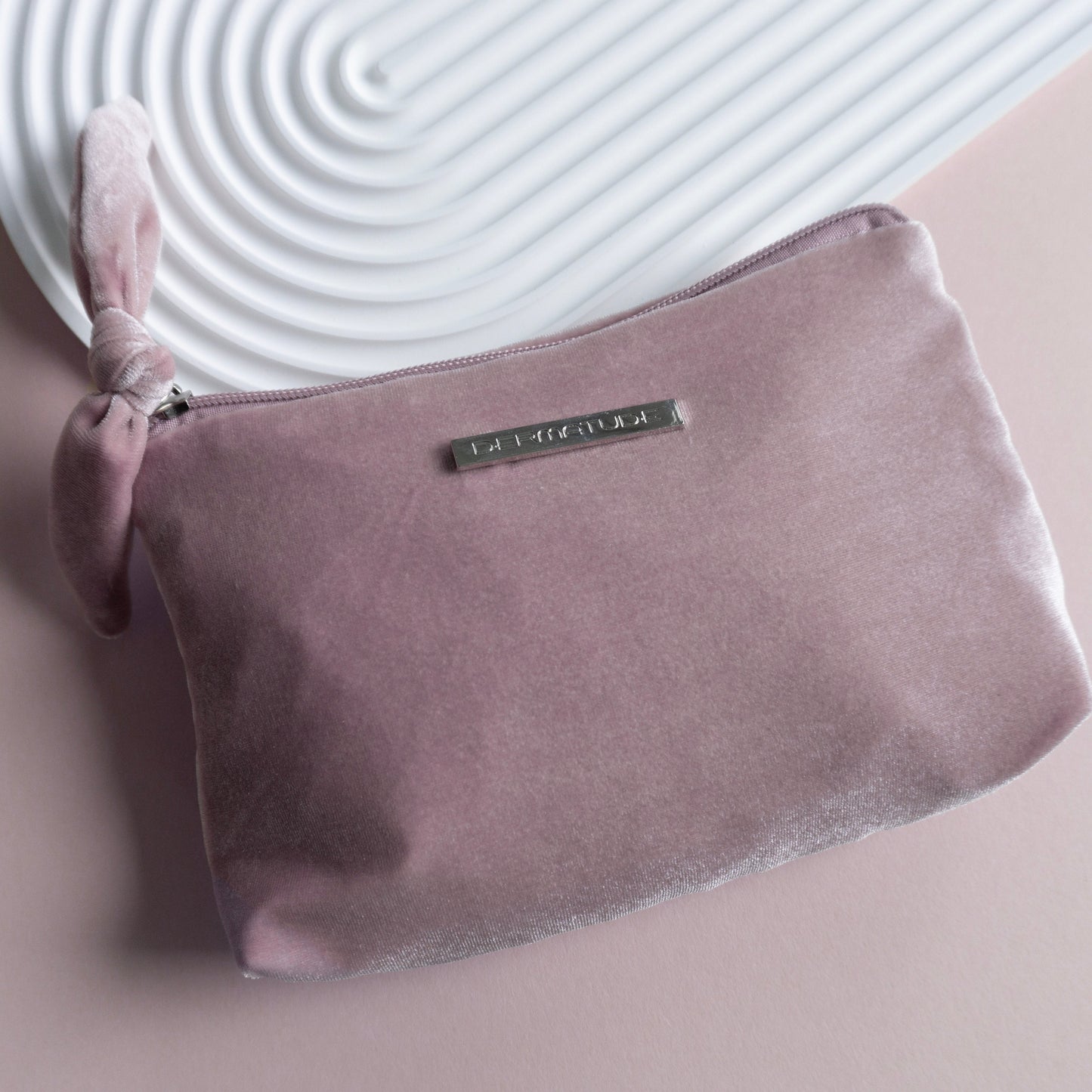 A pink velvet clutch with a silver pin that says DERMATUDE.